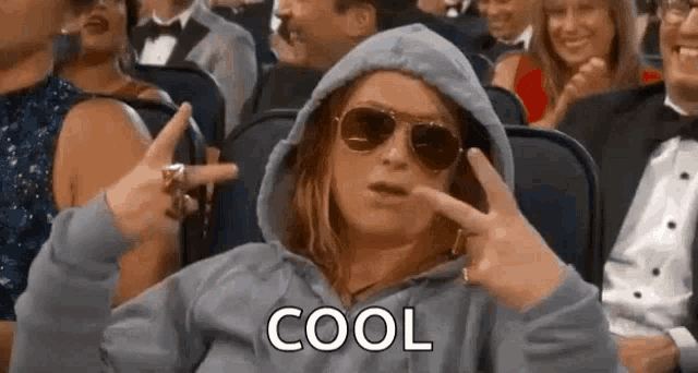 a woman wearing sunglasses and a hoodie is sitting in a crowd of people and making a peace sign .