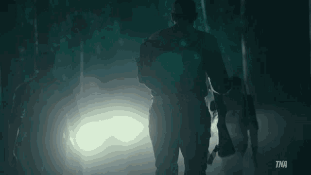 a man is walking through a dark forest with a tna logo on the bottom right