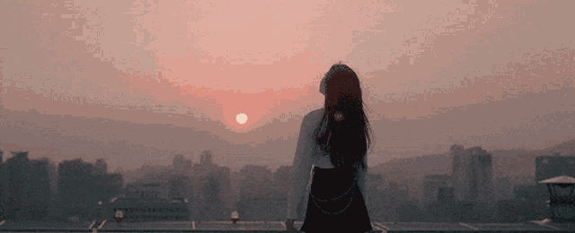 a woman stands on a balcony looking at the sunset over a city