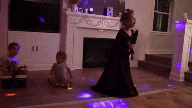a little girl in a black dress is singing into a microphone while two other children play drums in a living room .