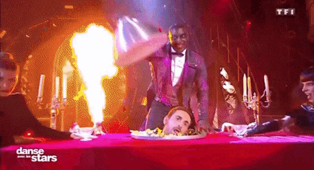 a man in a tuxedo is standing on a table with a plate of food and a fire behind him .