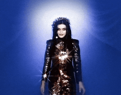 a woman in a black and gold dress and a crown is standing in front of a blue background .