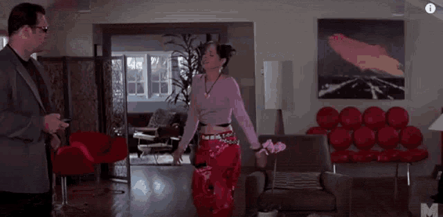 a man and a woman are standing in a living room . the woman is wearing a pink top and red pants .