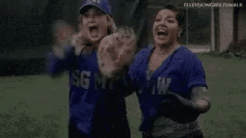 two women are standing next to each other on a baseball field and laughing .