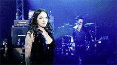 a woman in a black dress is standing on a stage in front of a man playing a guitar