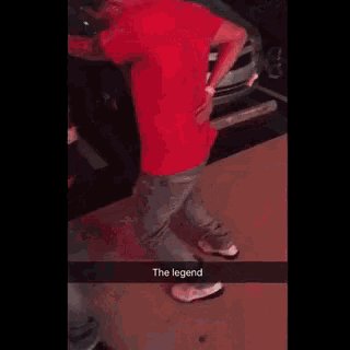 a man in a red shirt is standing in front of a car with his hands on his back .