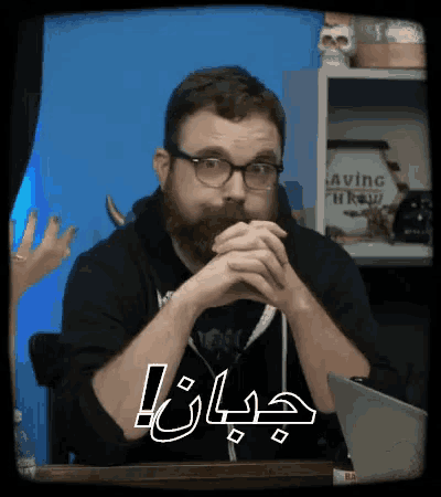 a man with glasses and a beard is sitting at a table with his hands folded in front of a sign that says saving