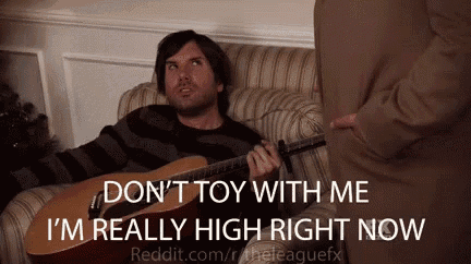 a man is sitting on a couch playing a guitar and talking to another man