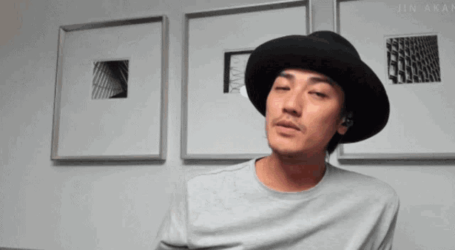 a man wearing a black hat is standing in front of a wall with pictures on it and the name jin akan on the bottom