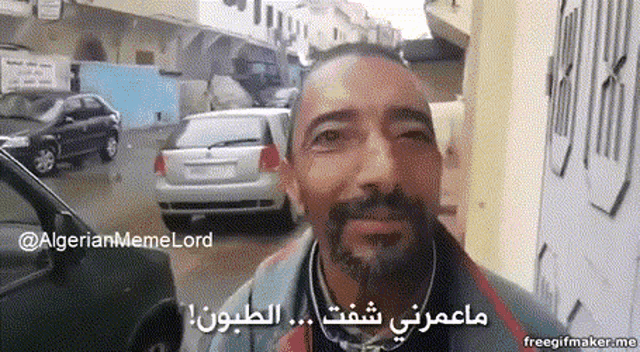 a man with a beard and glasses is smiling in front of cars and a sign that says " algerianmemelord "
