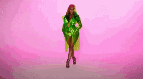 a woman is walking on a pink background wearing a green dress and a green trench coat .