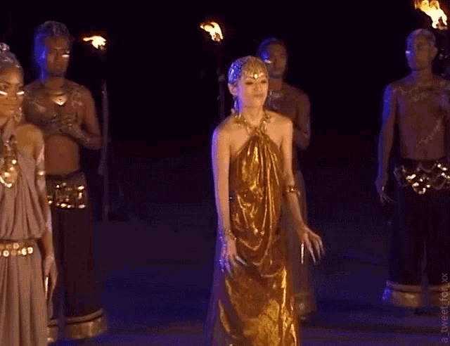 a woman in a gold dress stands in front of a group of people holding torches