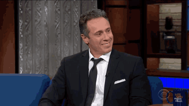 a man in a suit and tie is smiling while sitting on a couch in front of a sign that says late night