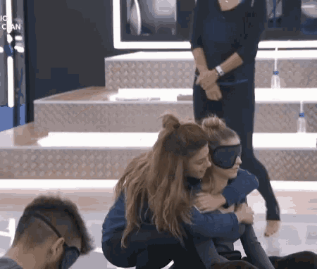 a woman wearing a blindfold is hugging another woman who is sitting on the floor .