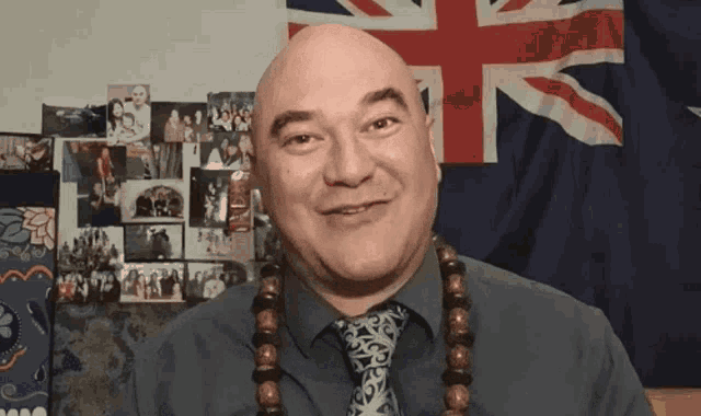a bald man wearing a tie and a necklace smiles in front of a british flag