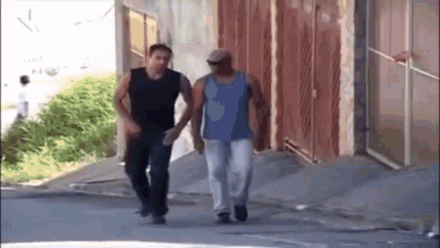 two men are walking down a street and one of them is wearing a blue tank top .