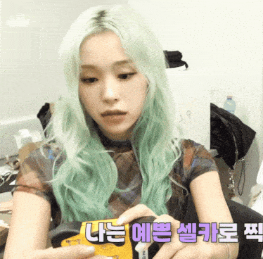 a woman with green hair is holding a yellow item with korean writing on the bottom