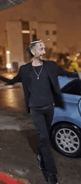 a man in a black jacket and black pants is standing next to a blue car