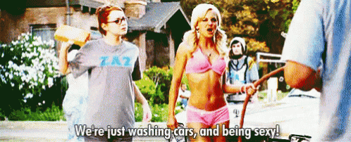 a woman in a bikini is standing next to a man holding a hose and talking about washing cars .