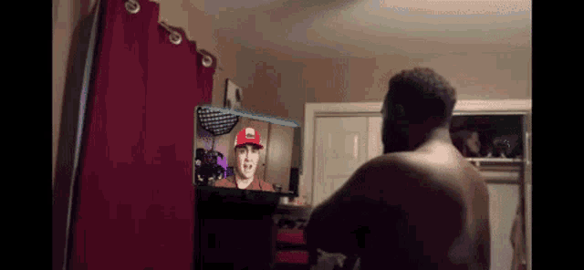a shirtless man is standing in front of a television with a man in a red hat on it .