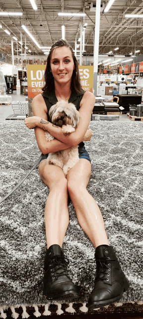 a woman sits on a rug holding a small dog in front of a sign that says loves on us