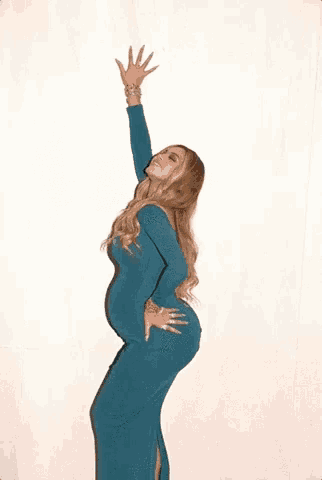 a pregnant woman in a blue dress is holding her belly and raising her hand .