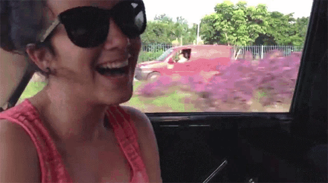 a woman wearing sunglasses and a pink tank top is laughing in a car