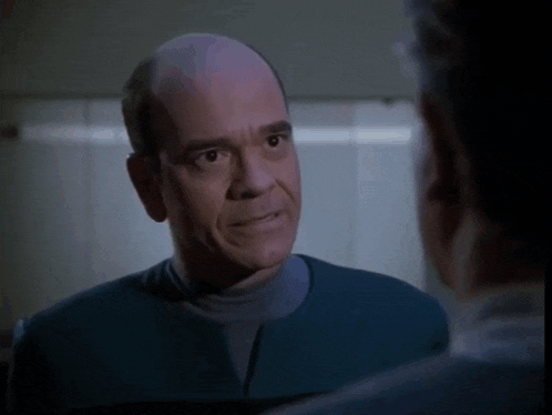 a bald man in a blue shirt talks to another man