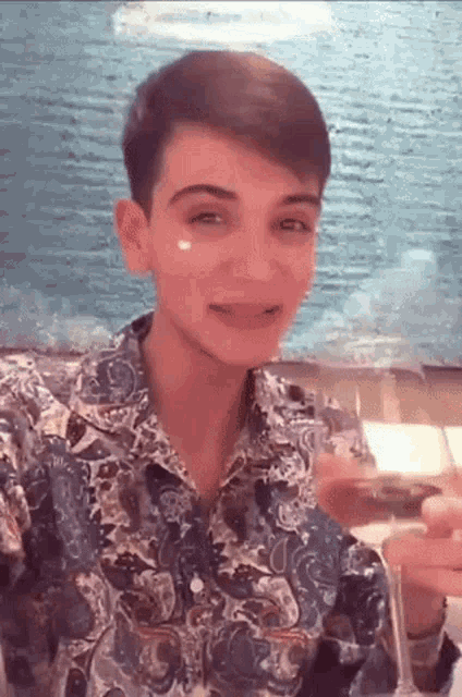a young man wearing a paisley shirt is holding a glass of wine and smiling .