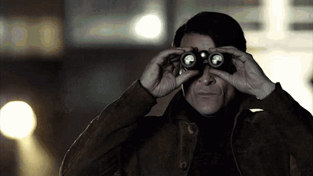 a man in a brown jacket looks through binoculars at something in the dark
