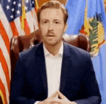 a man in a suit and white shirt is sitting in front of a flag .