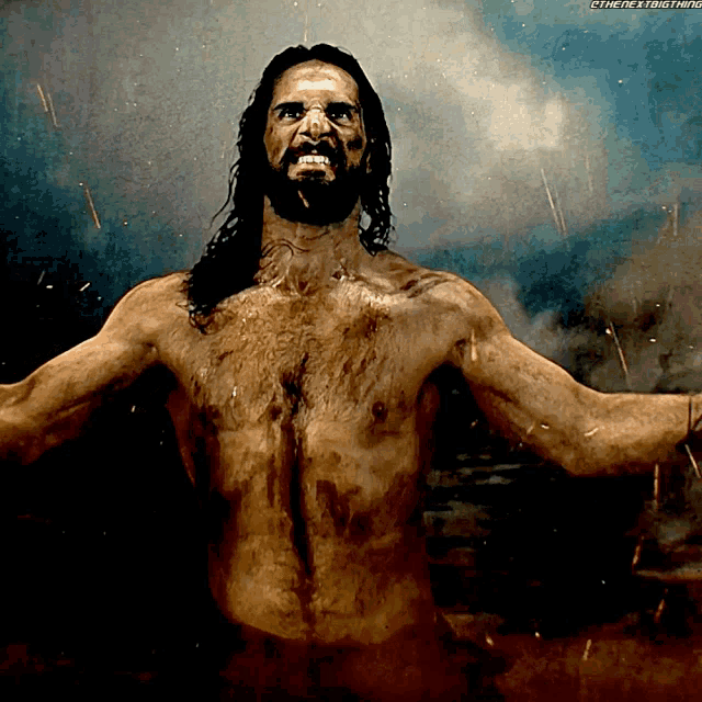 a shirtless man with long hair and a beard stands with his arms outstretched in front of a background that says the next thing
