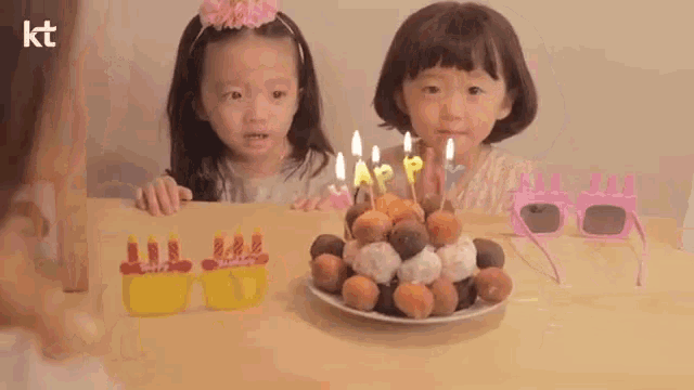 two little girls are sitting at a table with a birthday cake with candles on it .