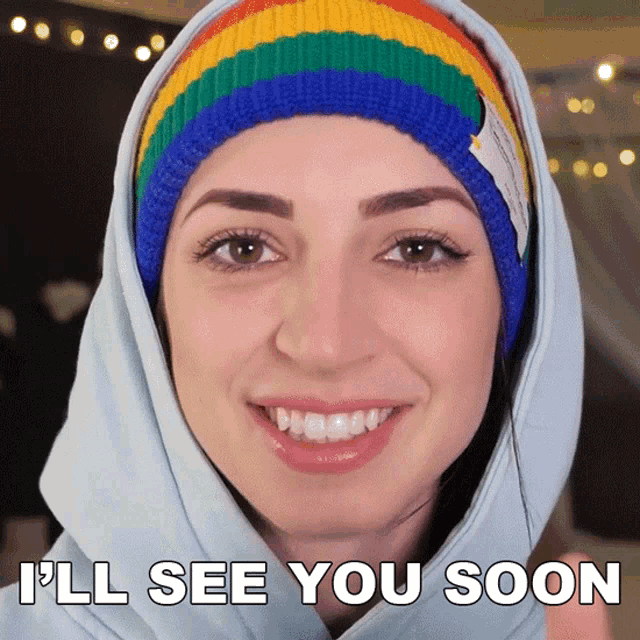 a woman wearing a rainbow colored beanie and a hoodie says i 'll see you soon