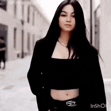 a woman wearing a black crop top and a black jacket is standing on a street .