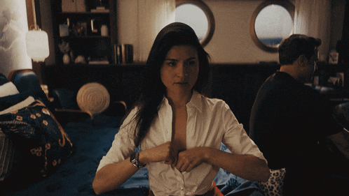 a woman in a white shirt is sitting on a bed and taking off her shirt
