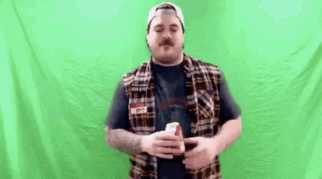 a man in a plaid vest and hat is holding a can of soda in front of a green screen .