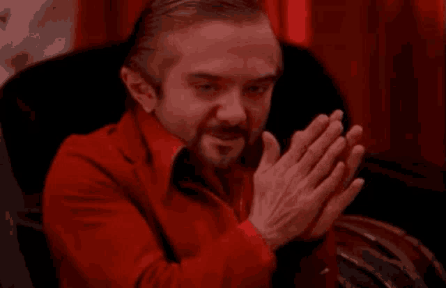 a man in a red suit is sitting in a chair with his hands folded .