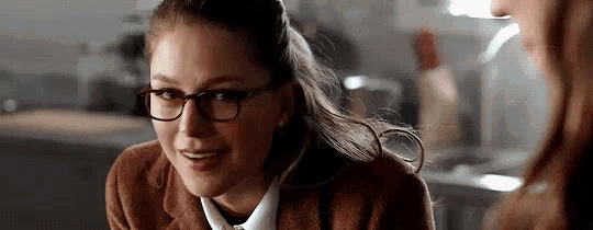 a woman wearing glasses and a brown coat is talking to another woman in a kitchen .