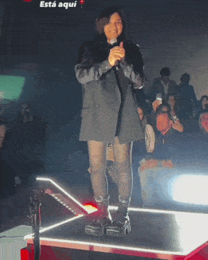 a woman stands on a stage with a microphone in front of a crowd and the word está aqui is on the bottom right