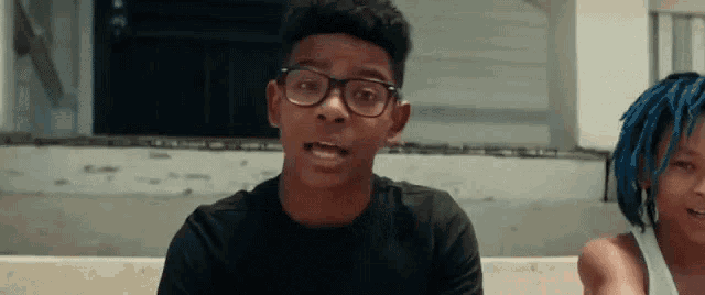 a boy wearing glasses and a girl with blue hair are sitting next to each other .