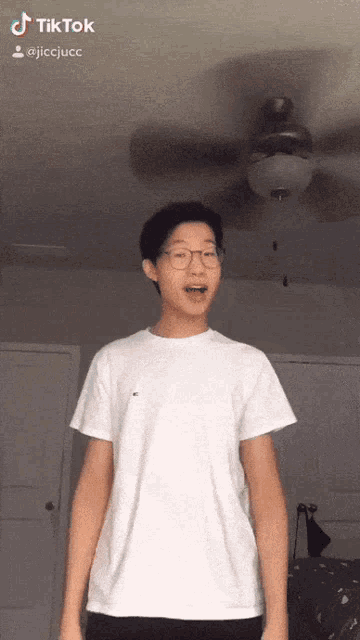 a young man wearing glasses and a white t-shirt is standing in front of a ceiling fan and says tiktok