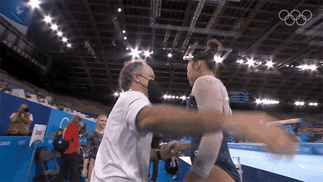 a man wearing a mask is hugging a female gymnast in a gym with the olympic rings in the background
