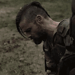 a man with a beard and mohawk is standing in the dirt in a field .