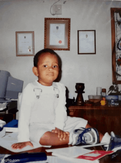 a young boy sits on a desk in front of a wall with framed certificates