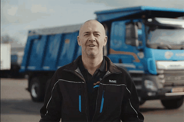 a man wearing a daf jacket stands in front of a blue dump truck