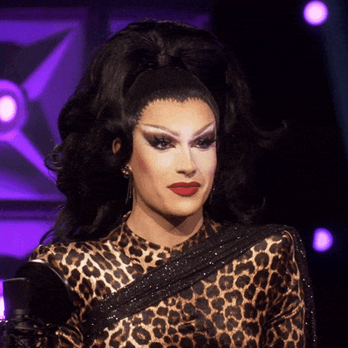 a drag queen in a leopard print top holds a mirror