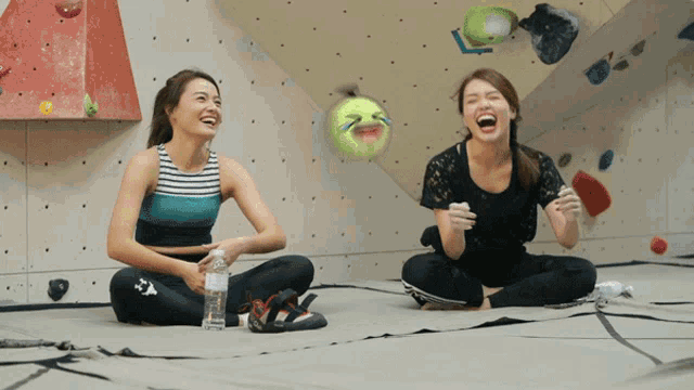 two women sit on the floor laughing with a ball that has a smiley face on it