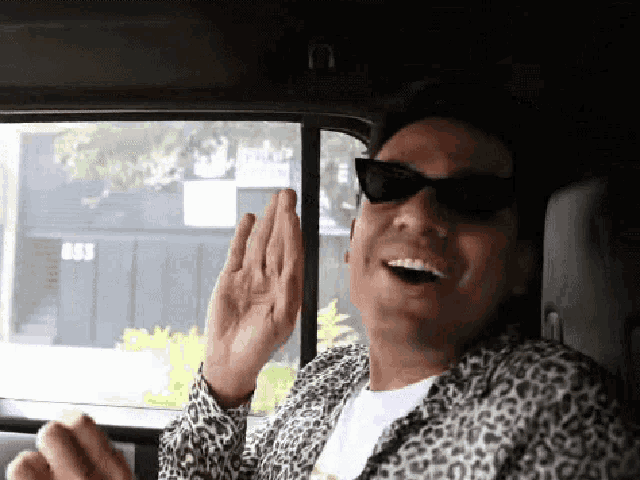 a man wearing sunglasses and a leopard print shirt is sitting in a car in front of a building with the number 853 on it