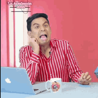 a man in a red and white striped shirt is sitting at a desk with a laptop and a coffee mug .